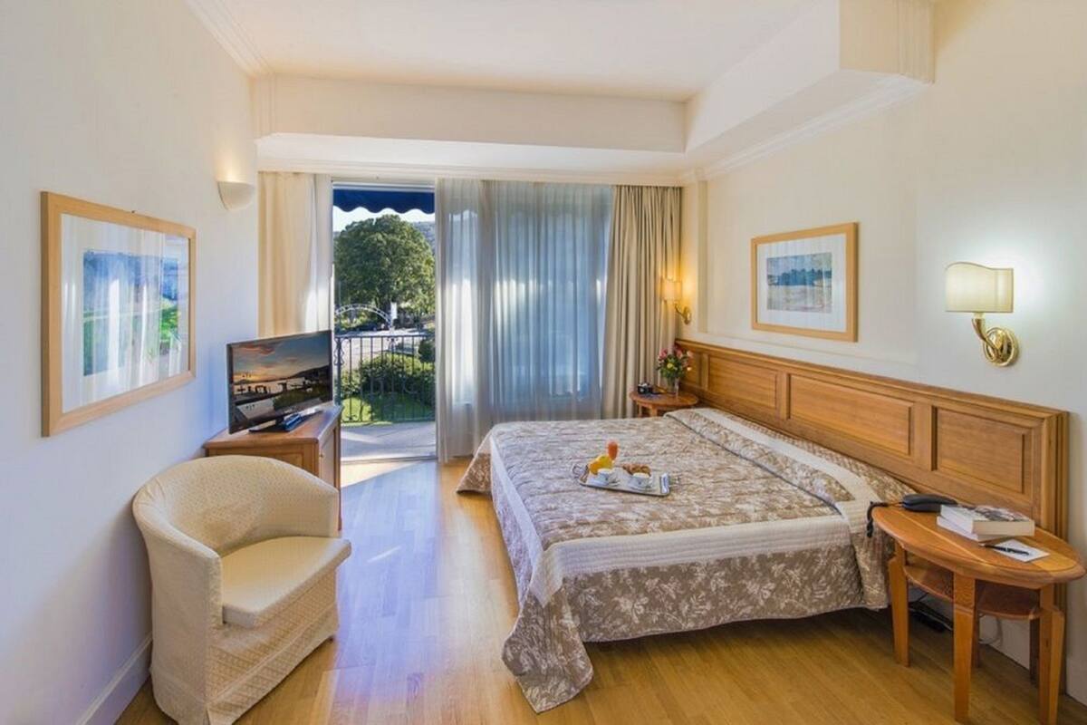 A hotel room with a bed, a chair, a TV, a bedside table, and a view of the outdoors.