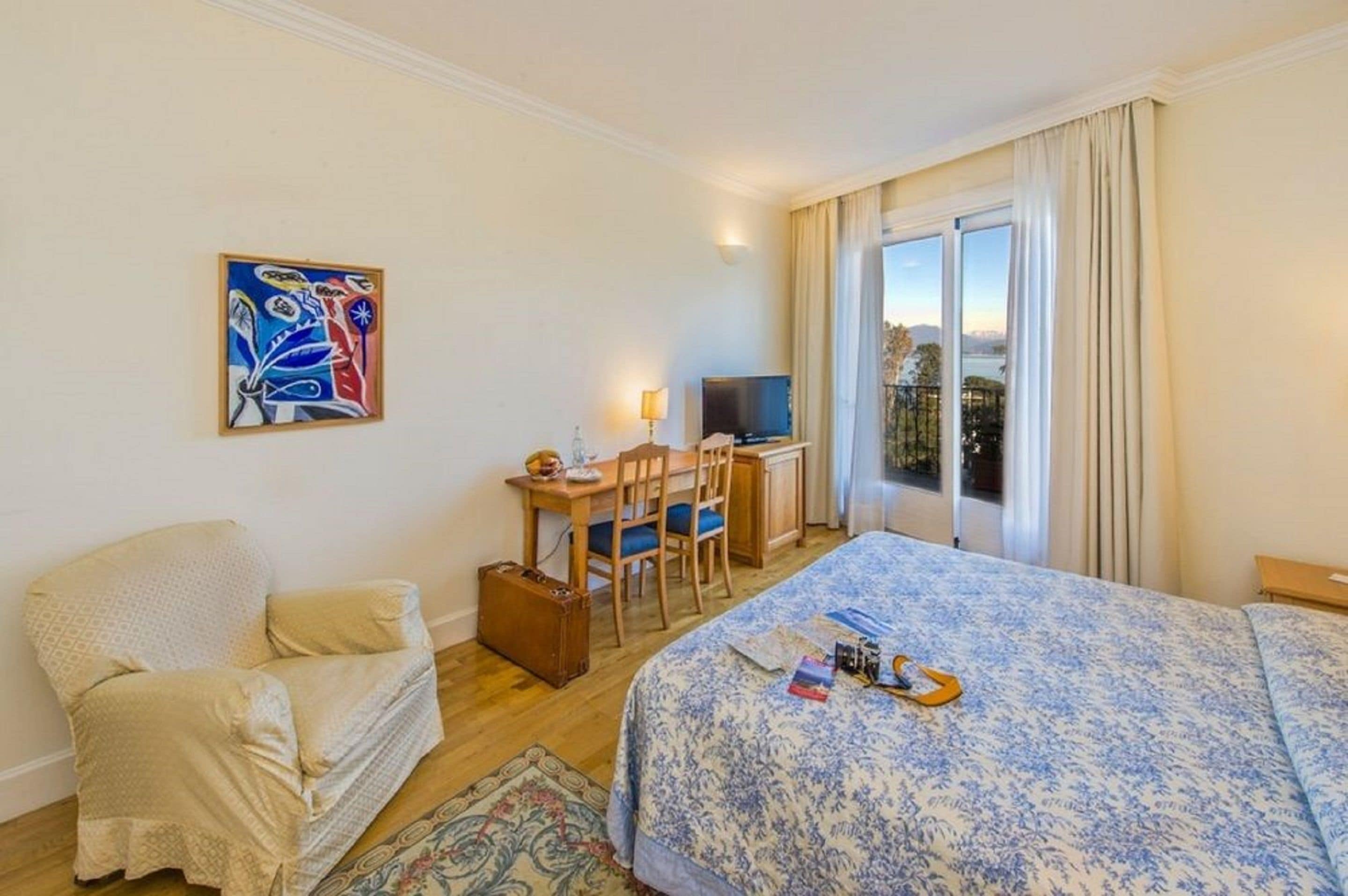 A hotel room with a bed, a desk with a TV, a chair, and a painting on the wall.