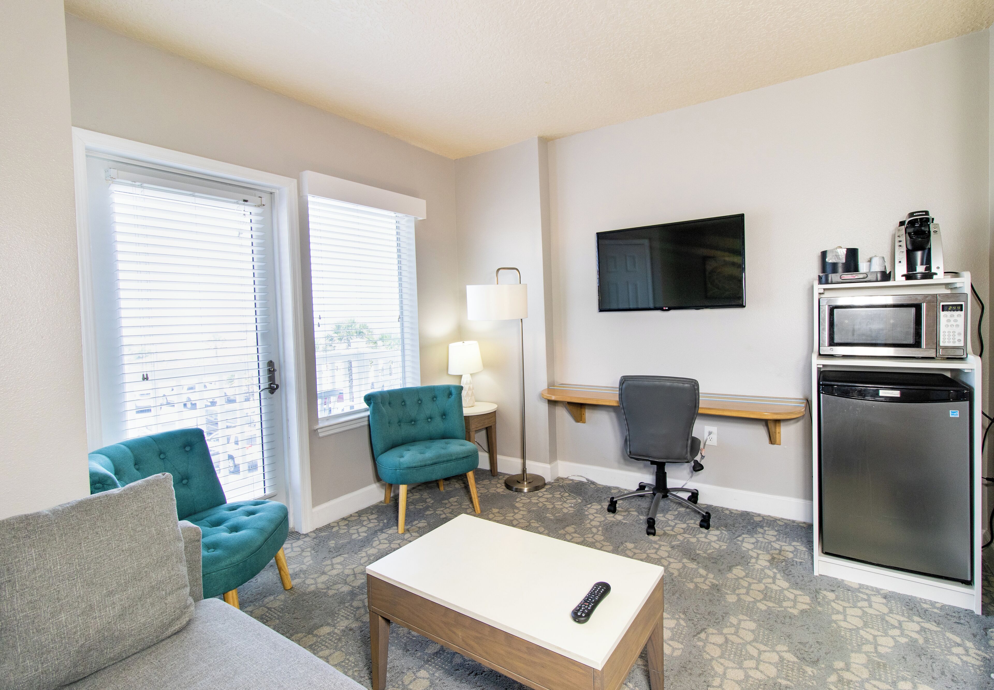 Habitación de hotel compacta con sofá, una pequeña mesa, un sillón azul verdoso, un escritorio con lámpara, televisor y una kitchenette con microondas y cafetera.