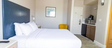 A hotel room with a bed, a desk, a chair, a microwave, and a door leading to a kitchenette.
