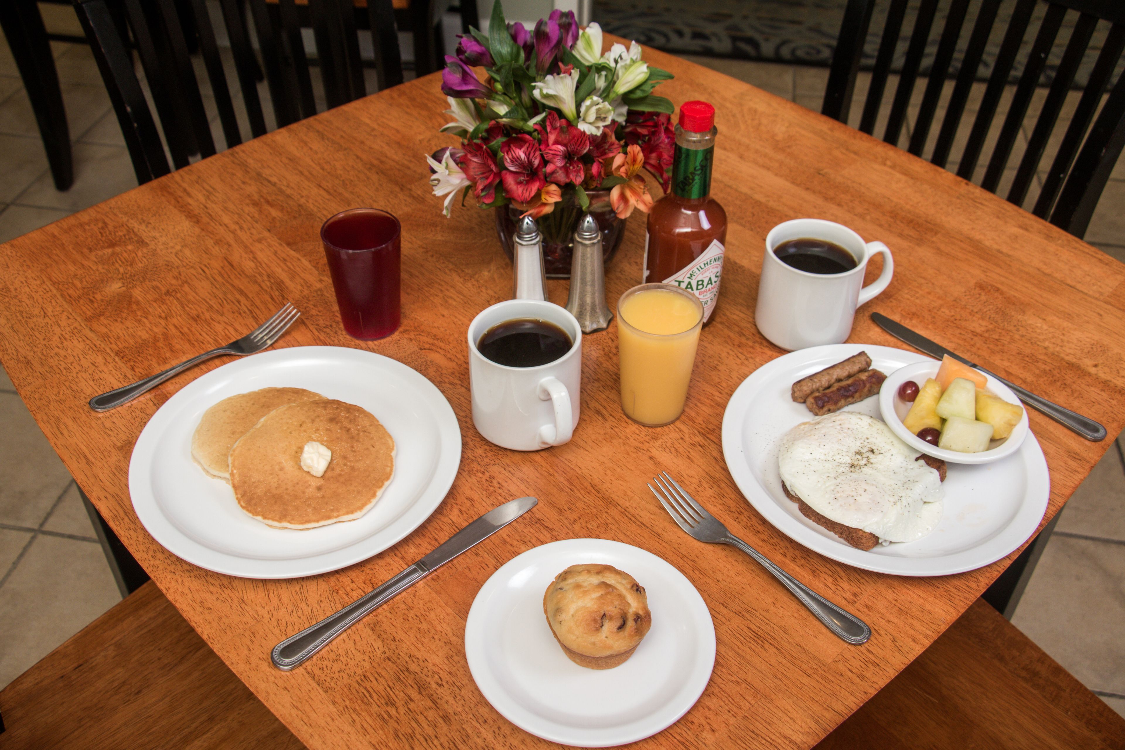 A wooden table with breakfast items including pancakes, eggs, and a muffin. There are salt and pepper shakers, a bottle of ketchup, a cup of coffee, and a glass of orange juice.