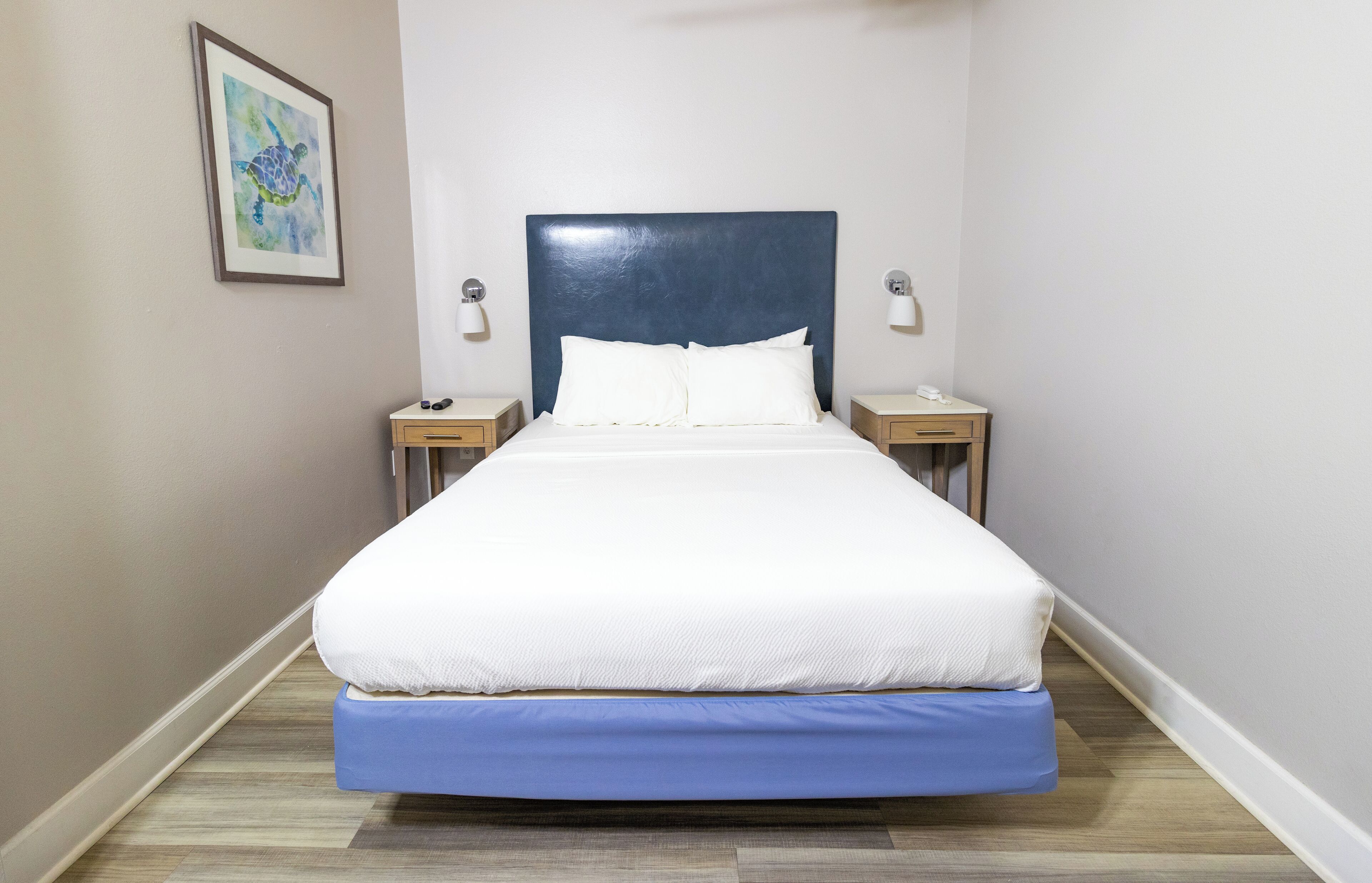 Een net opgemaakt bed met een blauwe matrasovertrek, witte kussens en een donkerblauwe hoofdbord. Aan beide zijden van het bed staan nachtkastjes met lampen.