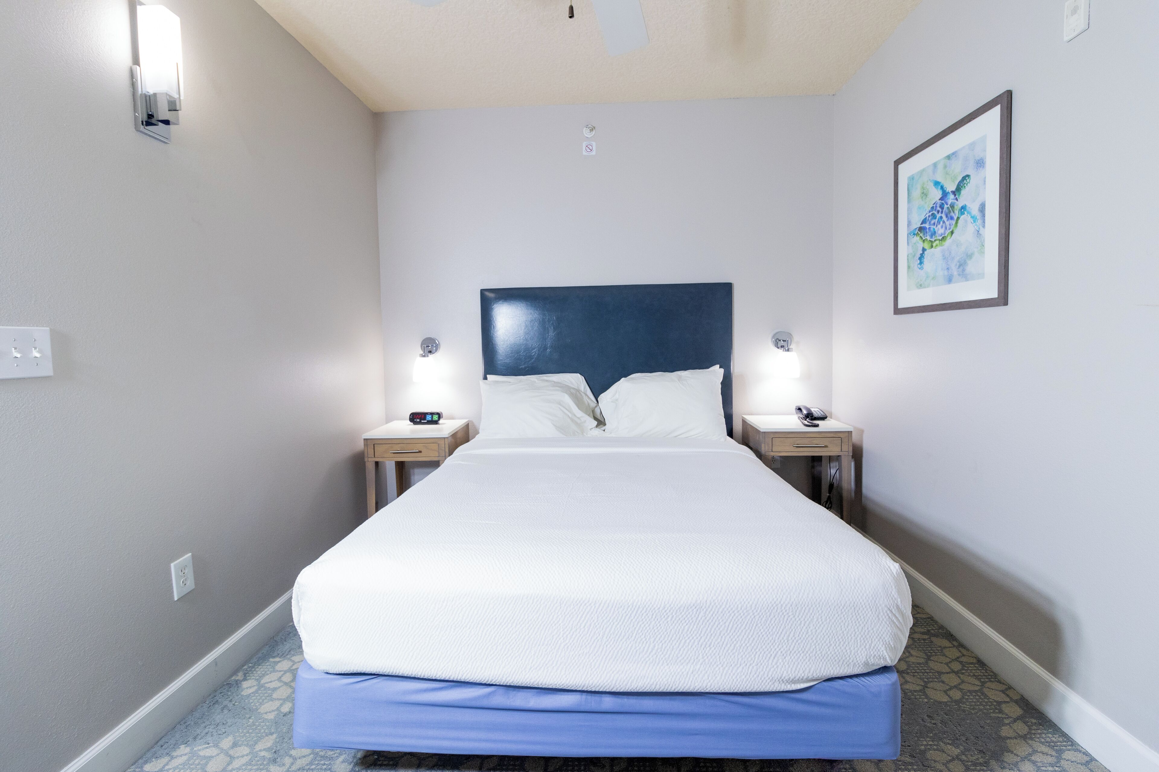 Ein Hotelzimmer mit einem großen Bett, Nachttischen, einem gerahmten Bild an der Wand und einem Lichtschalter.