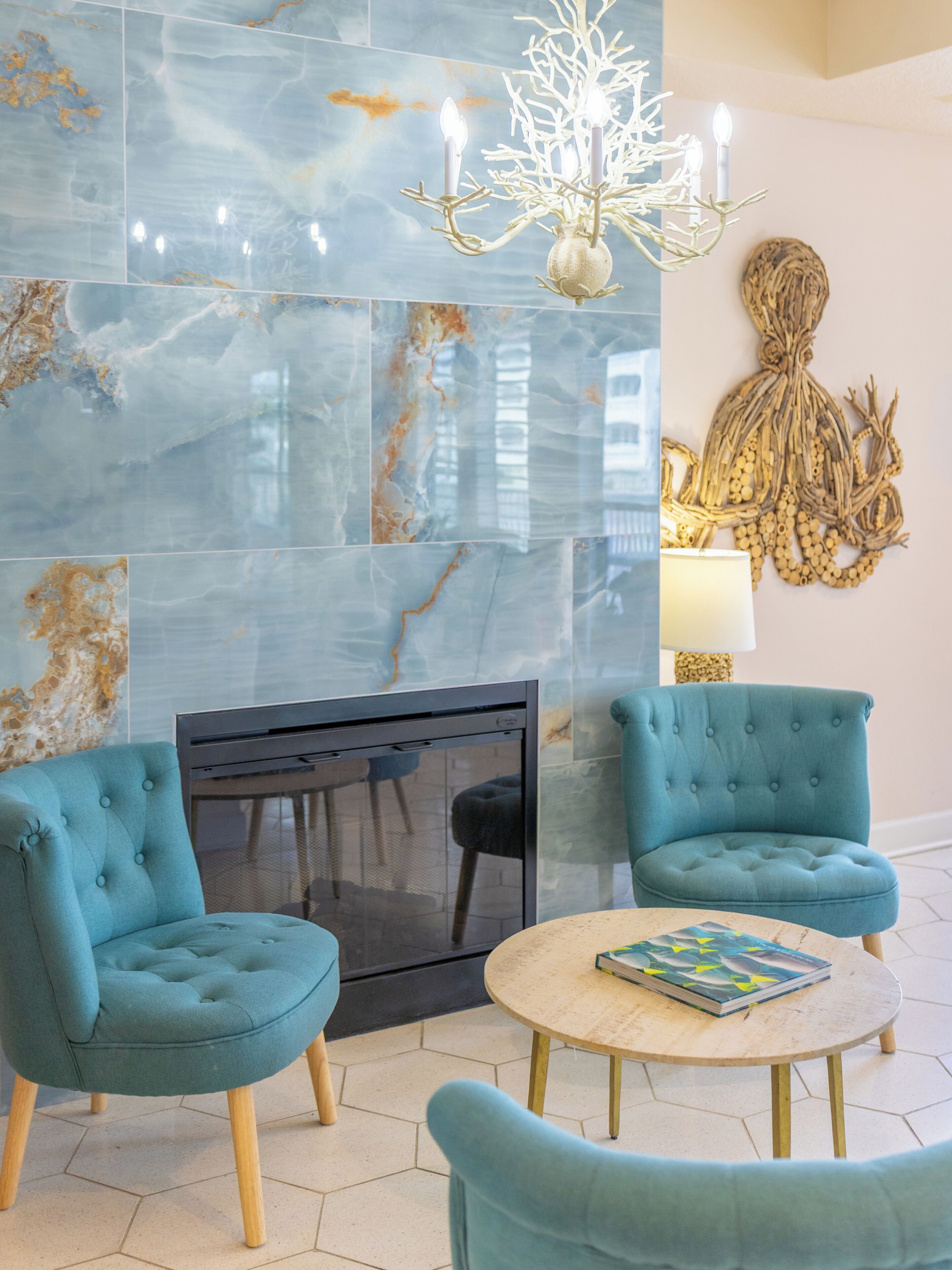 Un salon moderne avec une cheminée, deux fauteuils bleus, une table basse ronde et un lustre.