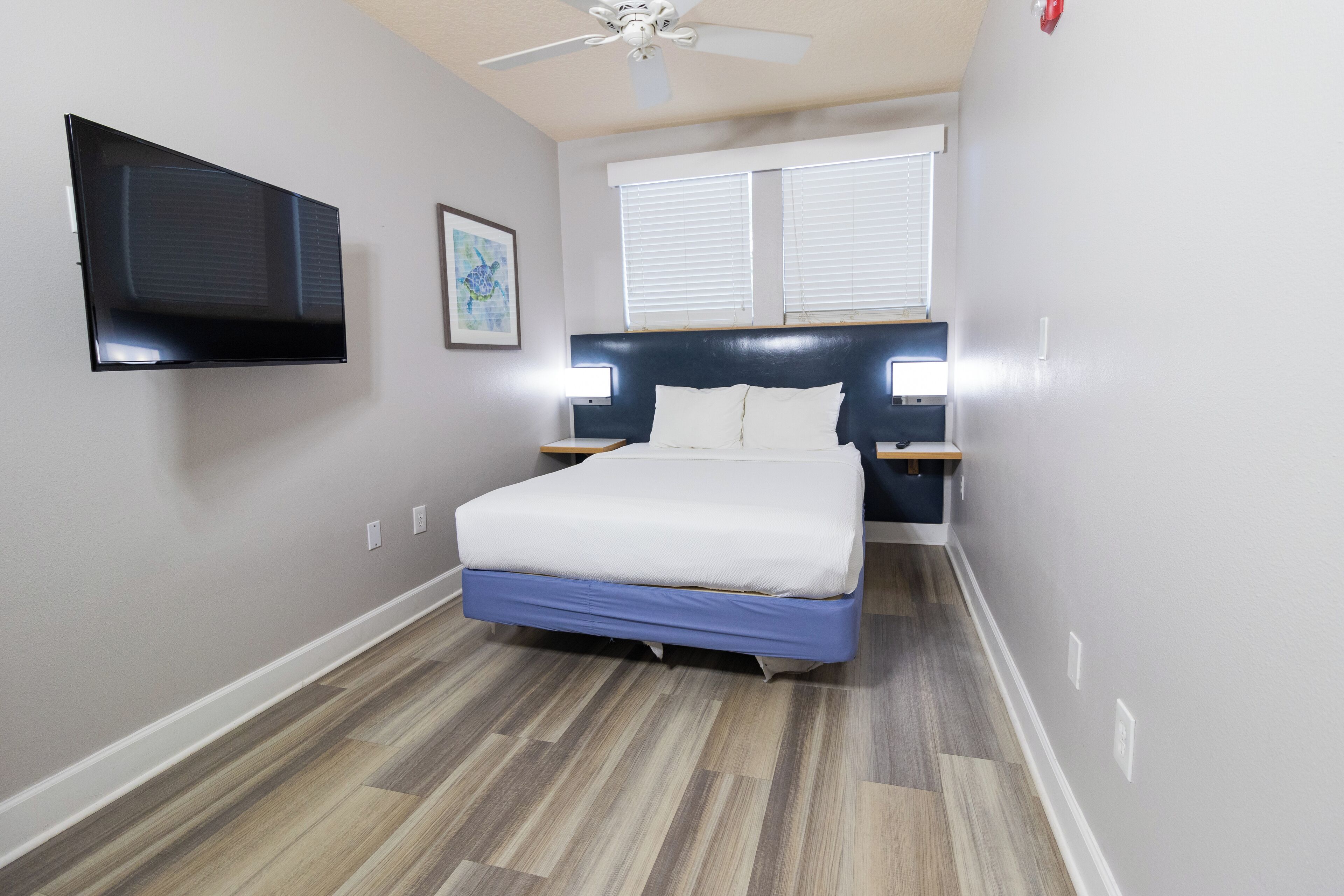 A bedroom with a bed, a ceiling fan, a TV mounted on the wall, and a window with blinds.