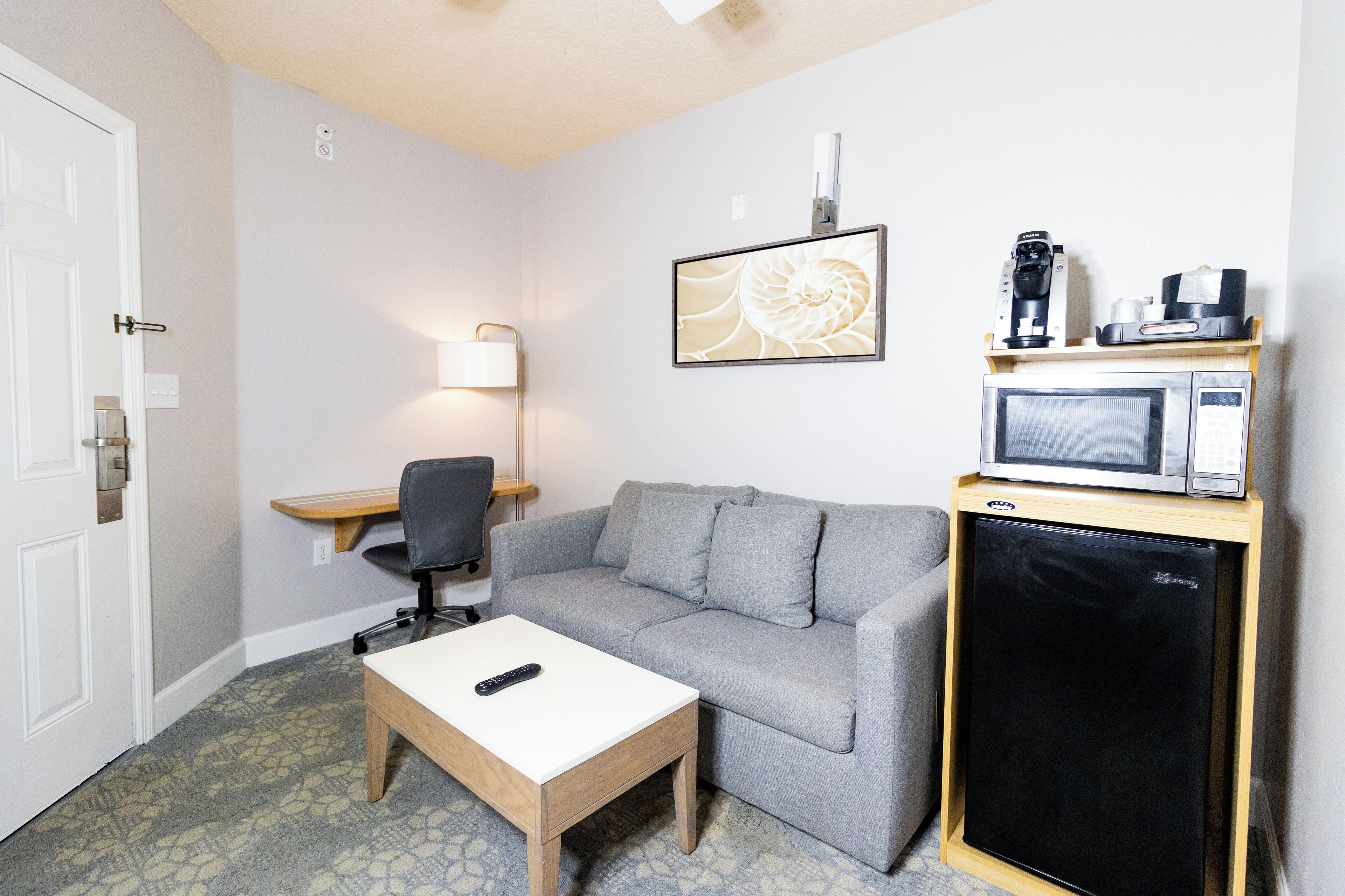 A compact hotel room with a grey sofa, a small wooden table, a black refrigerator, a microwave, and a coffee machine on a shelf.