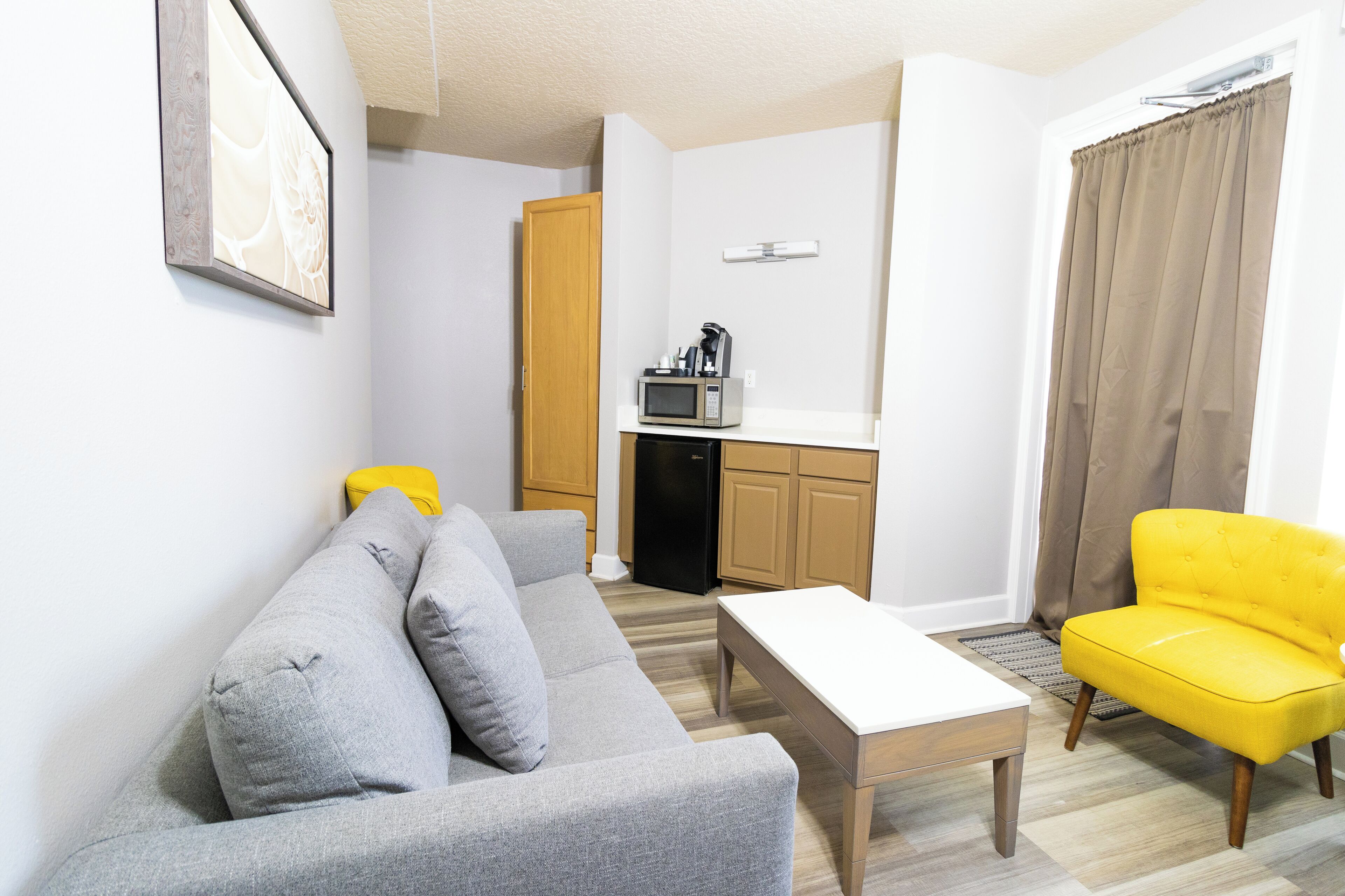 Een moderne woonkamer met een grijze bank, een gele fauteuil en een witte salontafel. In de achtergrond is een keuken te zien, met een magnetron en kastjes.