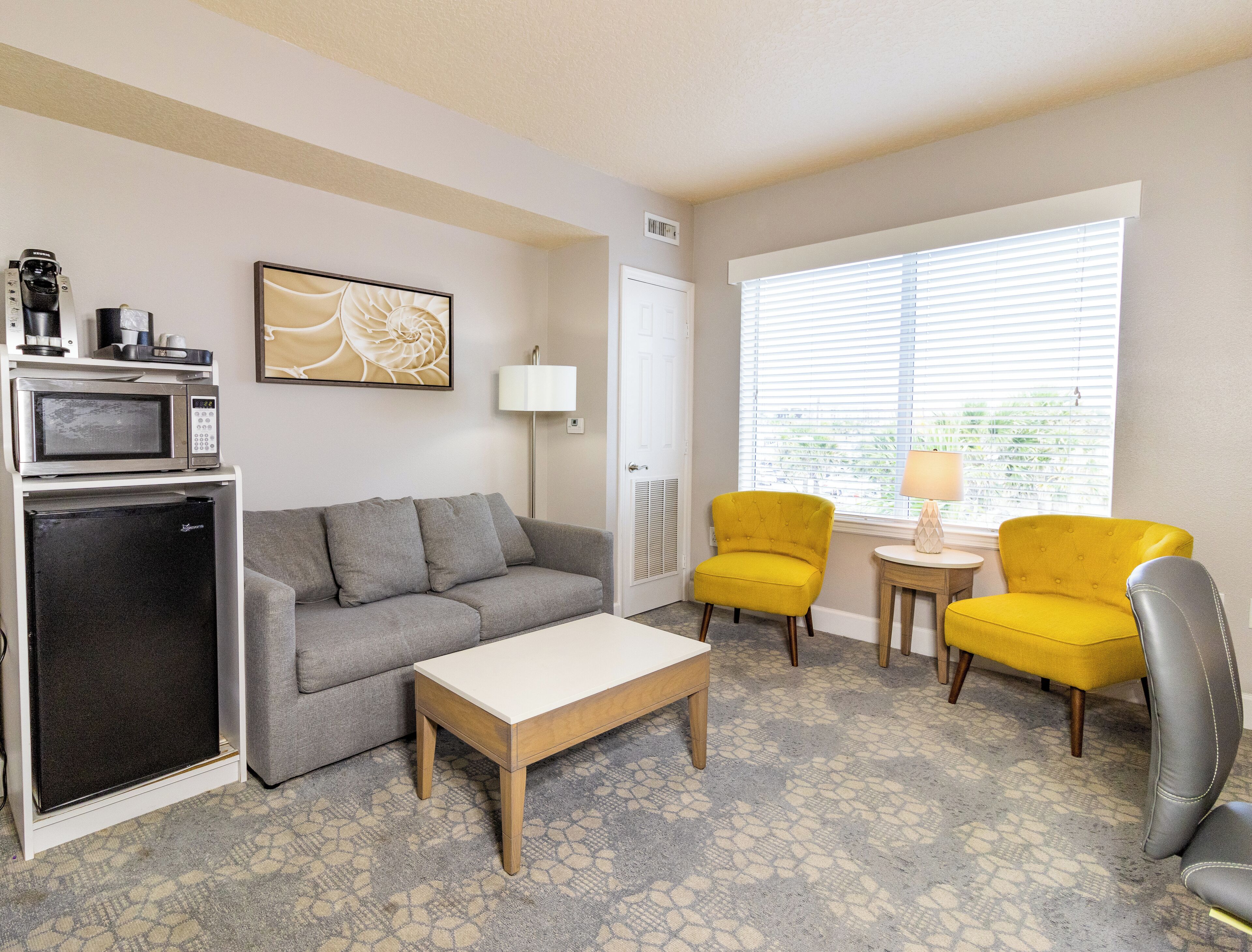 A compact living space with a grey sofa, a small wooden coffee table, a microwave, a mini fridge, two yellow armchairs, a small side table with a lamp, and a large window with blinds.