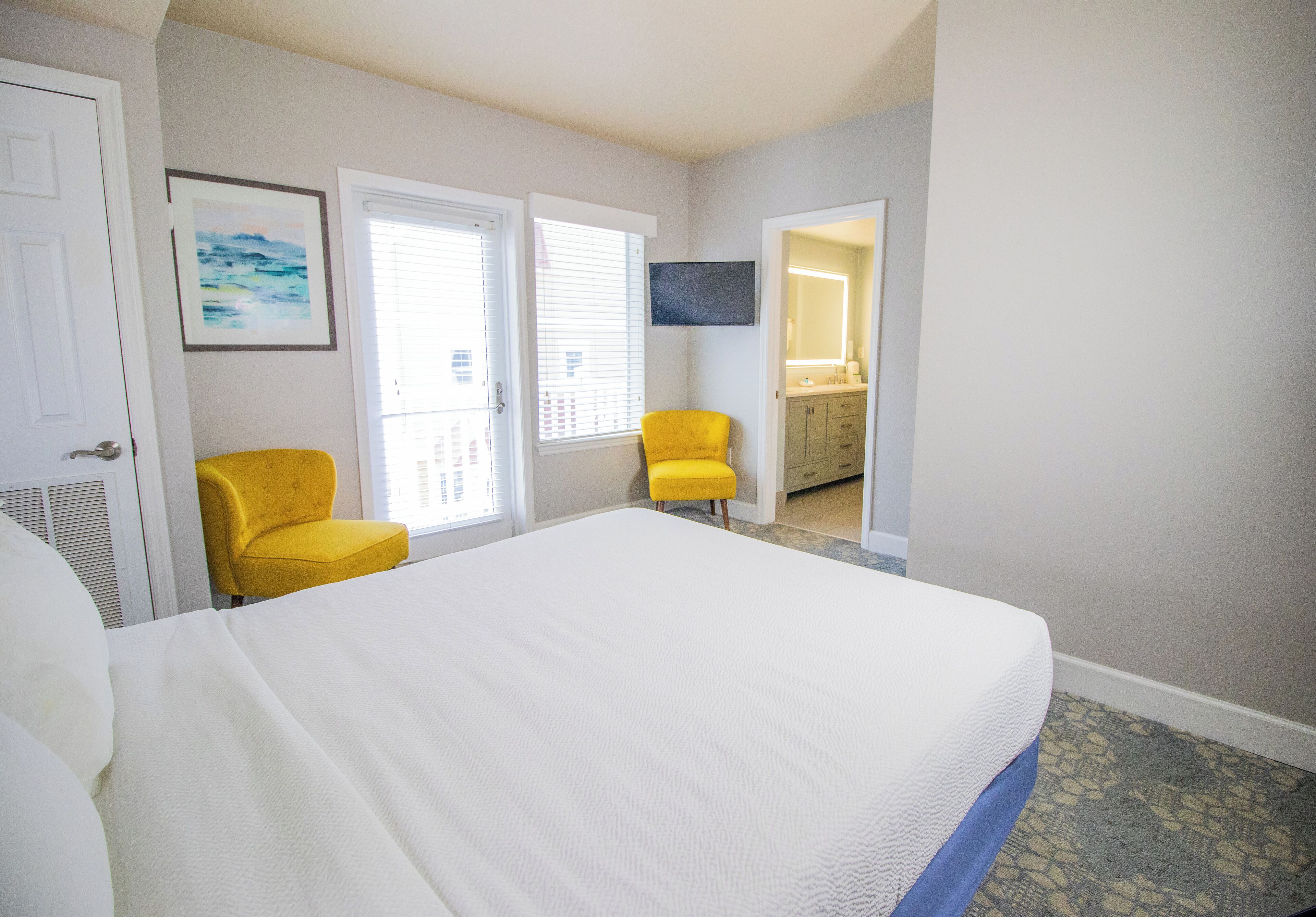 A bedroom with a large bed, two yellow chairs, a TV, and a bathroom visible through an open door.