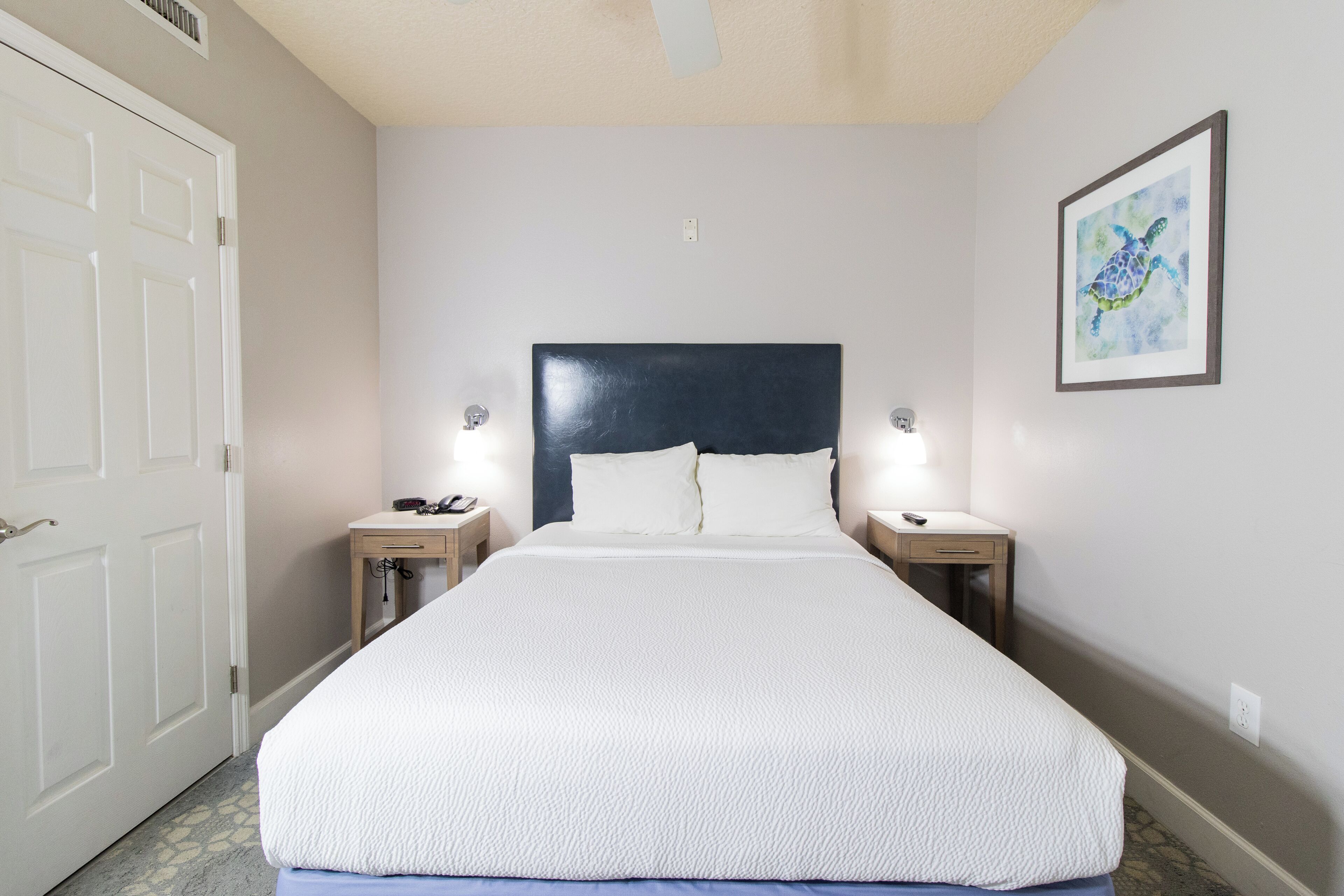 Un letto rifatto con un copriletto bianco e cuscini, due comodini con lampade, un quadro appeso al muro e una porta bianca.