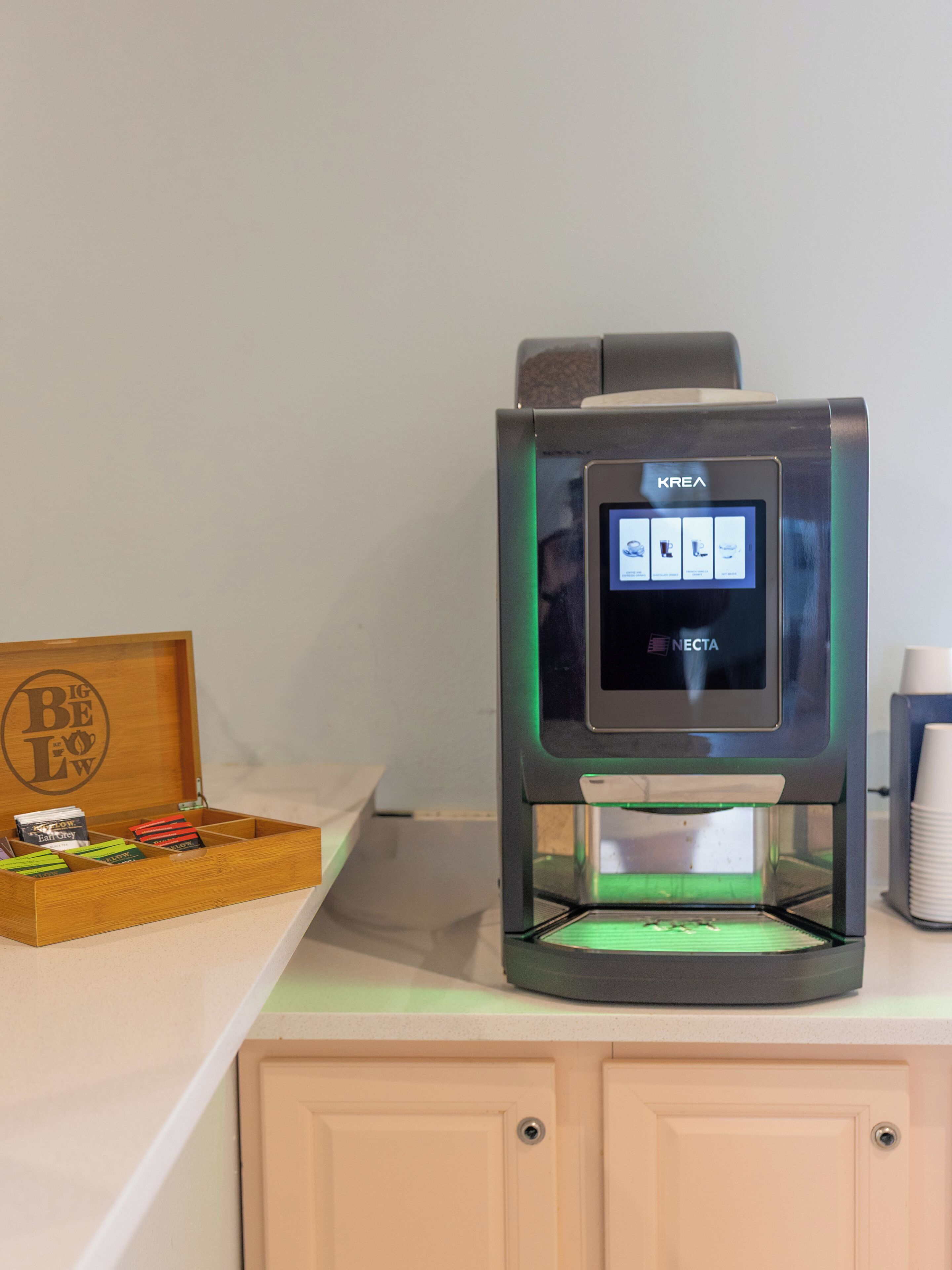 A coffee machine with a digital display, a wooden box with tea bags, and a stack of paper cups.
