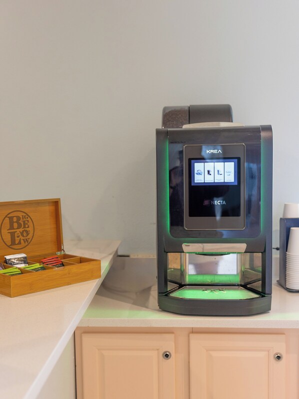Une machine à café dotée d’un écran numérique, une boîte en bois contenant des sachets de thé et une pile de gobelets en papier.