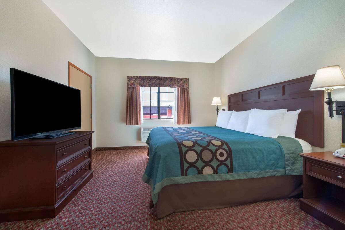 A hotel room with a bed, a television on a dresser, a window with curtains, and a nightstand with a lamp.