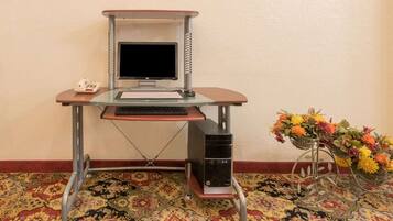 A glass desk with a computer monitor, keyboard, and phone. A desktop computer is on a stand beside the desk. A decorative metal stand with flowers is next to the desk.