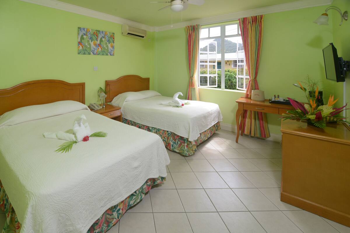 A hotel room with two beds, a desk, a chair, a window with curtains, a plant, and a painting on the wall.