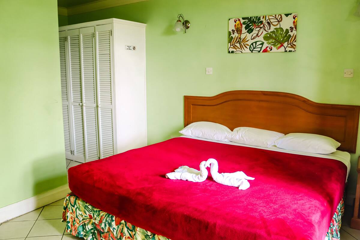 A bed with a red bedspread and two swan-shaped towels, a wooden headboard, and a wall-mounted light fixture.