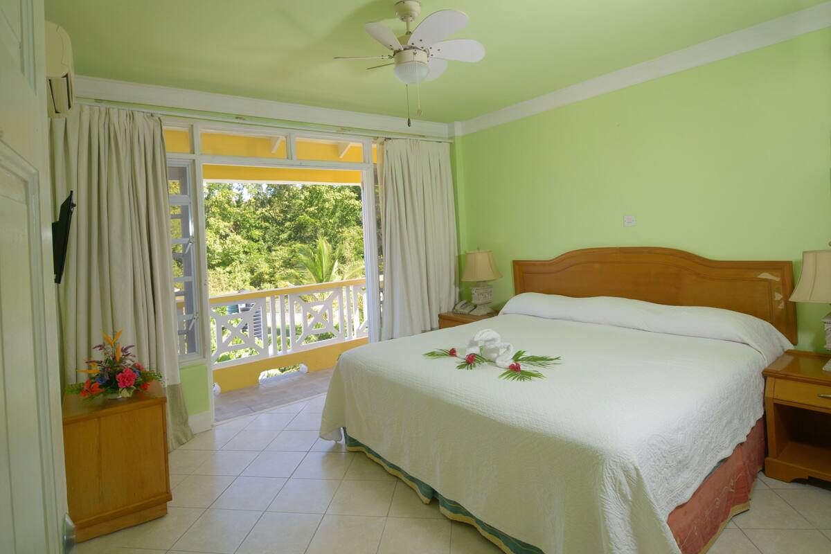 A bedroom with a bed, a ceiling fan, a nightstand, a lamp, and a view of greenery outside.