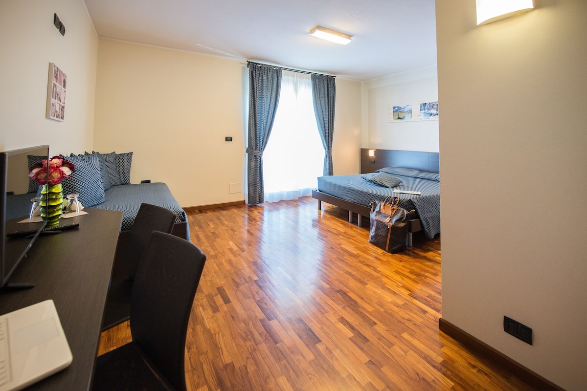 A hotel room with a wooden floor, a bed with a blue bedspread, a desk with a computer, and a chair.