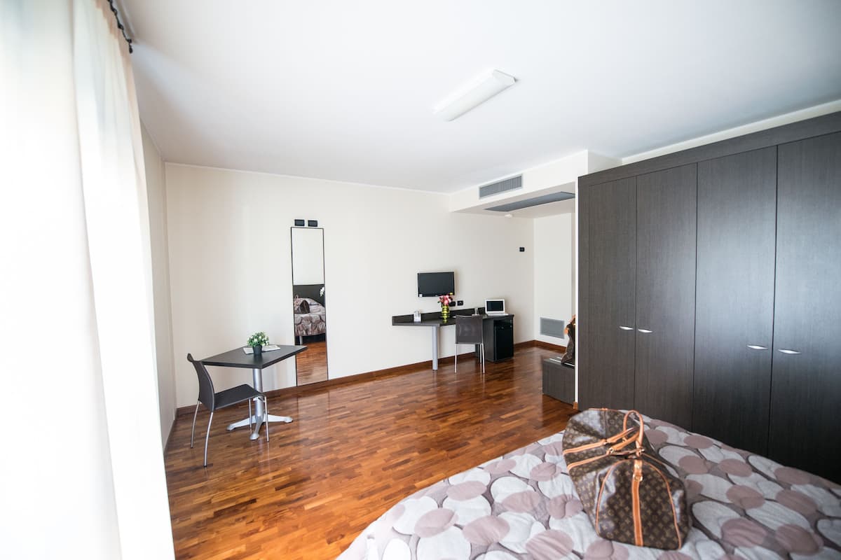 A modern hotel room with a wooden floor, a bed with a patterned bedspread, a desk with a computer, a chair, a wardrobe, and a mirror on the wall.