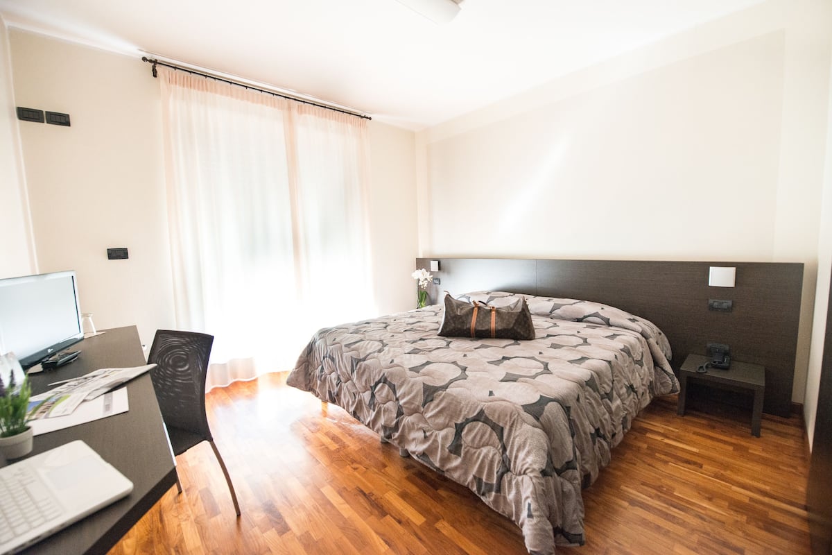 A bedroom with a bed, a desk with a laptop, a chair, a bedside table, and a nightstand.