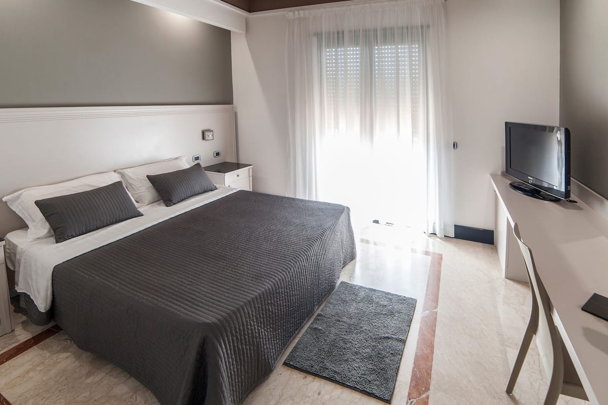 Una habitación de hotel moderna con una cama grande, un escritorio con televisor y una ventana con cortinas translúcidas.