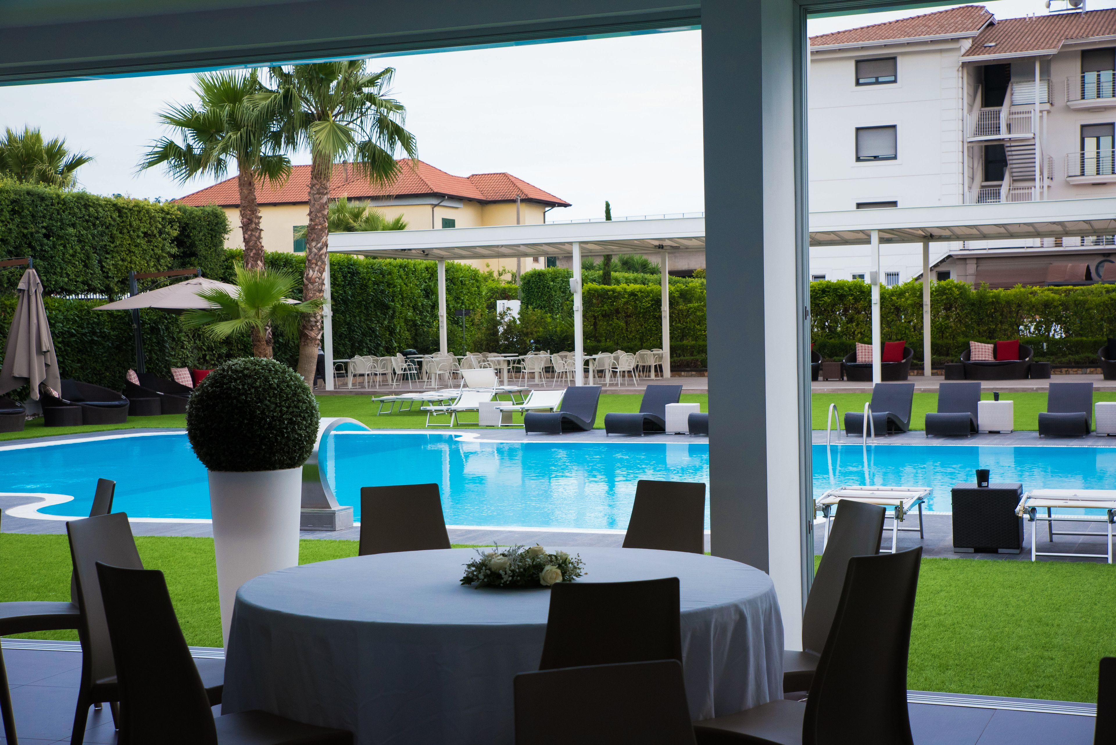 Un espace aménagé au bord de la piscine, avec des chaises longues et une table, une vue sur un bâtiment doté d’un toit rouge et une pelouse bien entretenue.
