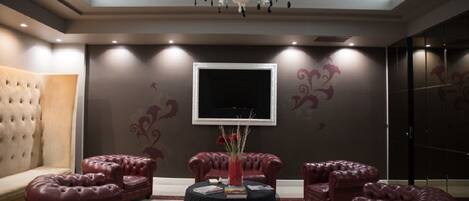 A luxurious room with a large sofa, multiple leather ottomans, a round table with a vase, and a flat-screen TV on the wall.