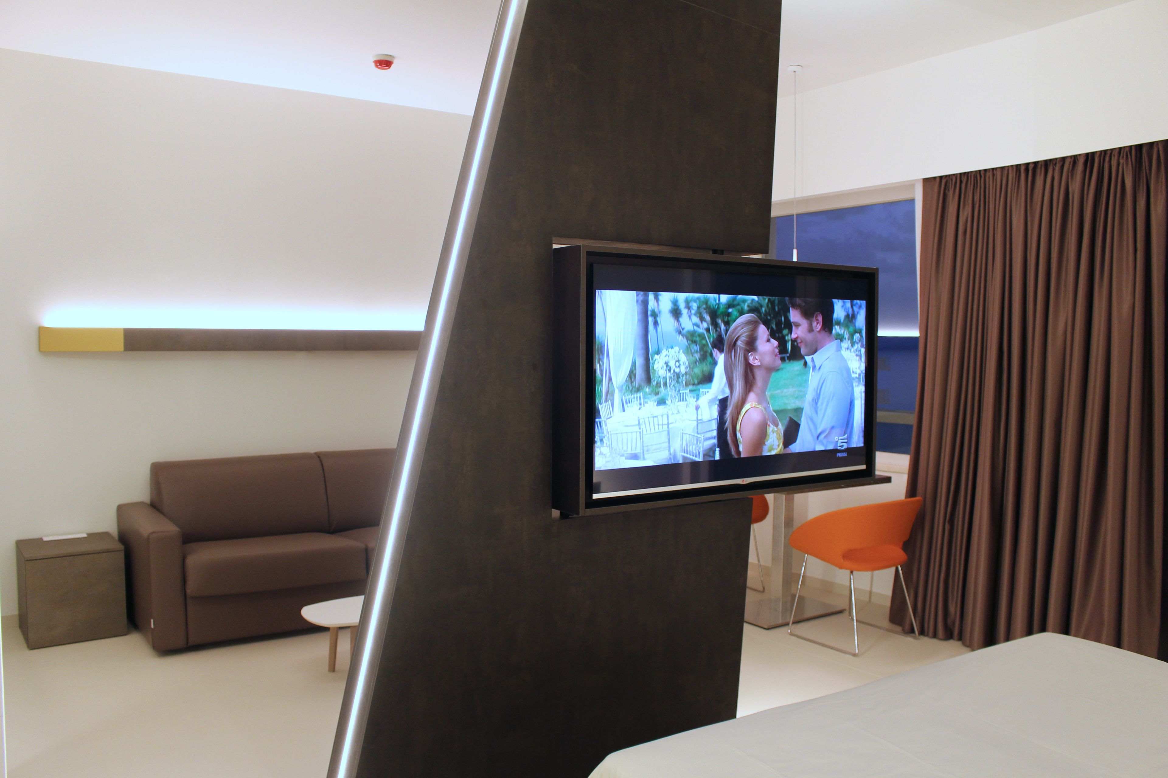 A modern hotel room with a flat-screen TV mounted on a wall, a brown sofa, a small round table, and an orange chair.