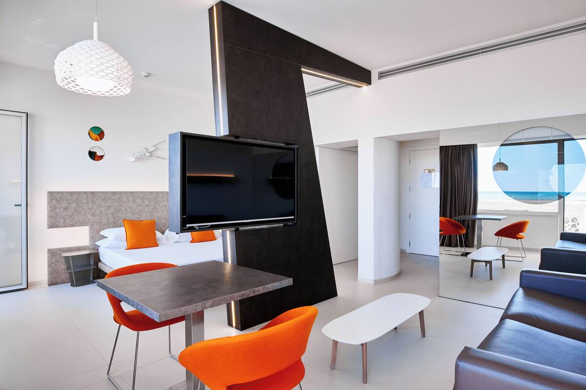 A modern hotel room with a dining area, a large TV, orange chairs, a sofa, and a view of the sea.