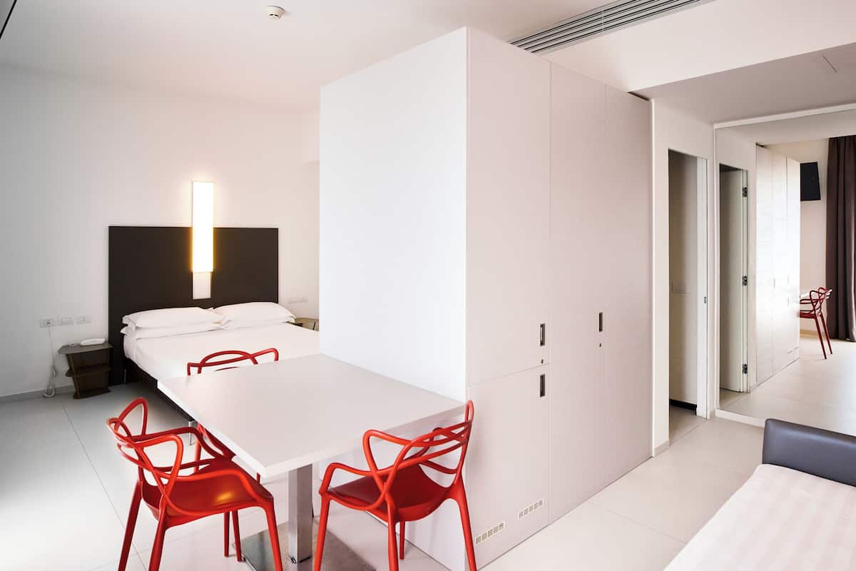 A modern hotel room with a white bed, a white desk, and red chairs. There is a wardrobe with a mirror and a bedside table with a lamp.