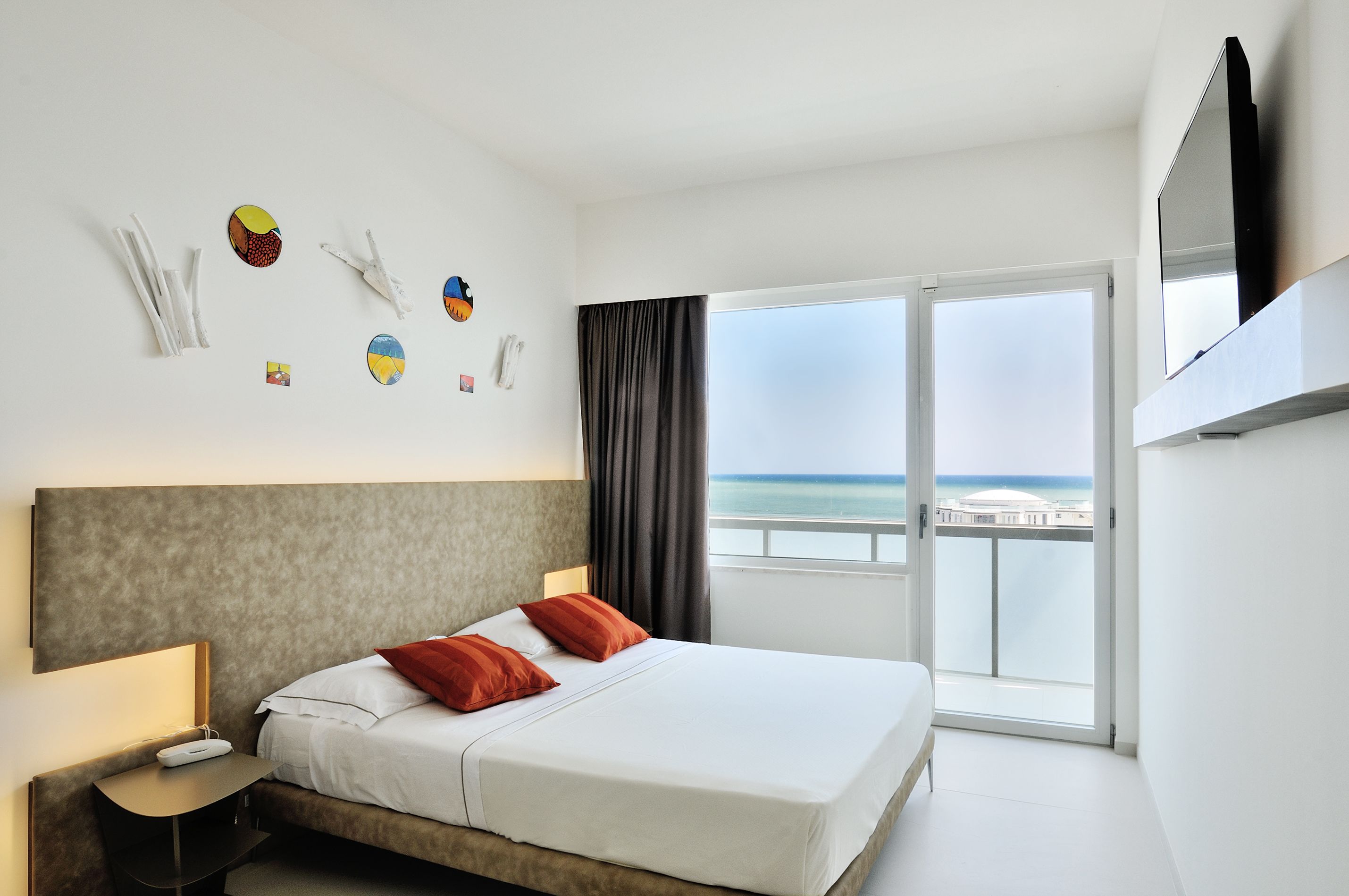 Habitación de hotel con cama, mesita de noche, televisor de pantalla plana y una ventana grande con vista al océano.