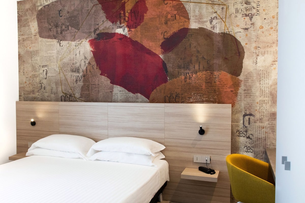 A modern hotel room with a large bed, a wooden headboard, a bedside table with a lamp, and a unique abstract wall art piece.