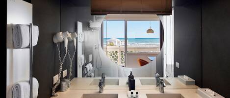 A modern bathroom with dual sinks, a large mirror, and a view of the ocean.