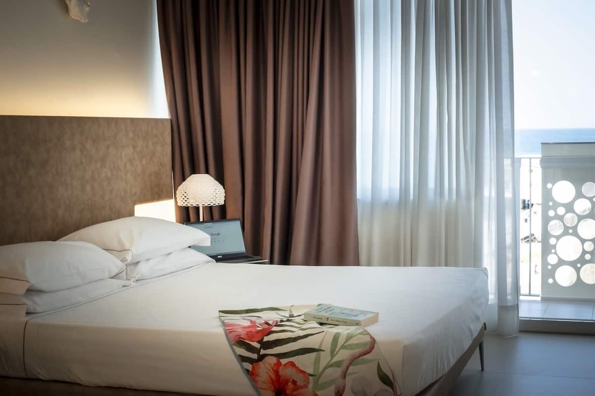 A hotel room with a bed, a laptop on a nightstand, and a view of the sea through a window with curtains.