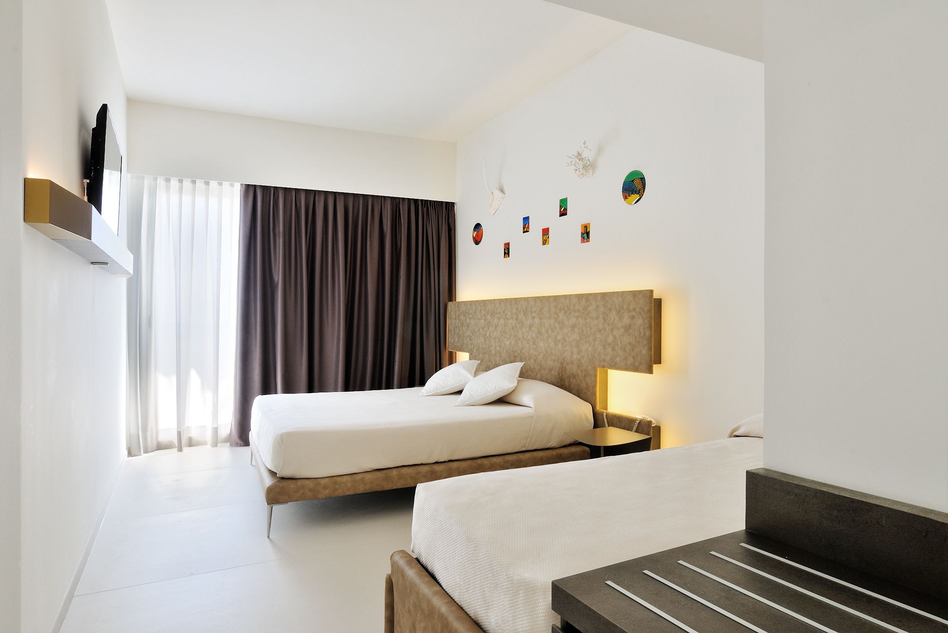 A hotel room with a bed, a bedside table, a TV mounted on the wall, and a window with curtains.