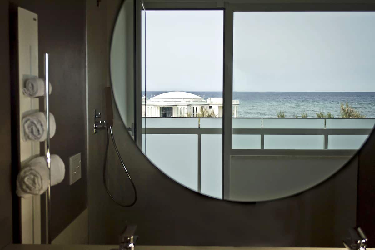 A bathroom with a large round mirror, a shower, and a view of the sea and a building through the window.