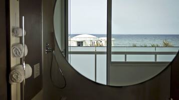 Un bagno con un ampio specchio rotondo, una doccia e vista sul mare e su un edificio dalla finestra.