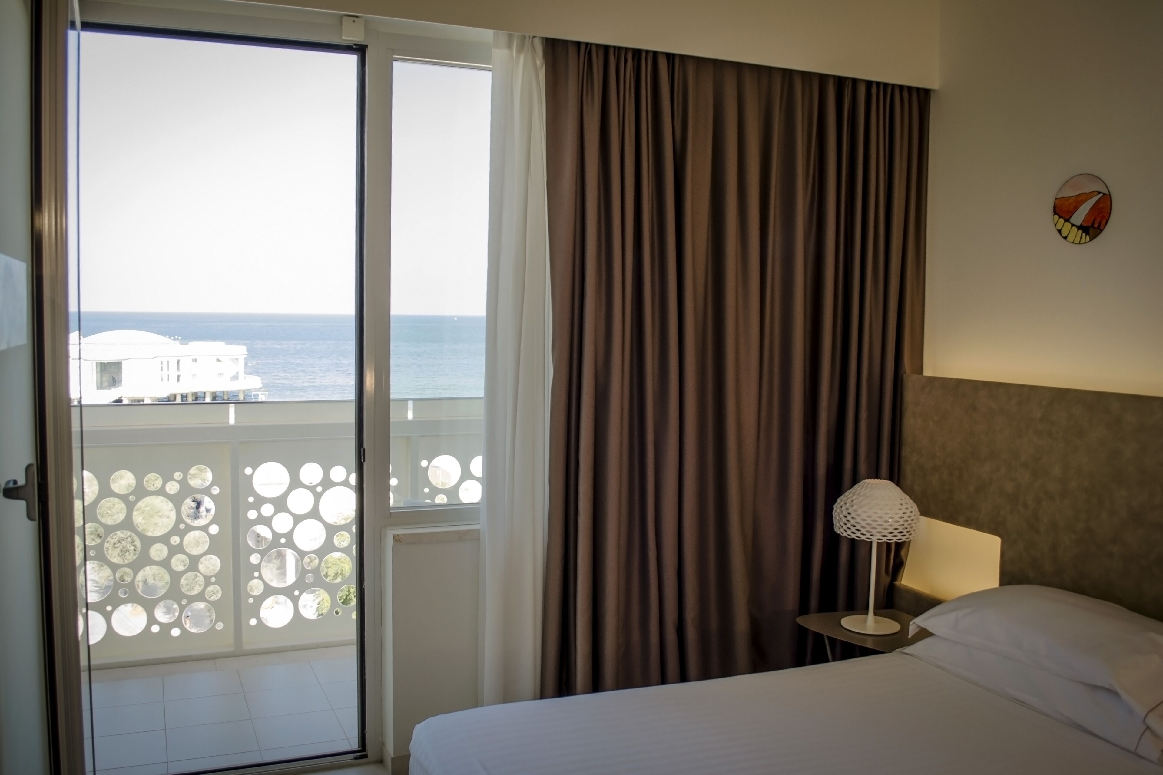 Habitación de hotel con cama, mesita de noche, lámpara y vista al mar a través de un ventanal con cortinas.