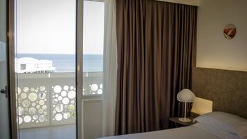 Habitación de hotel con cama, mesita de noche, lámpara y vista al mar a través de un ventanal con cortinas.