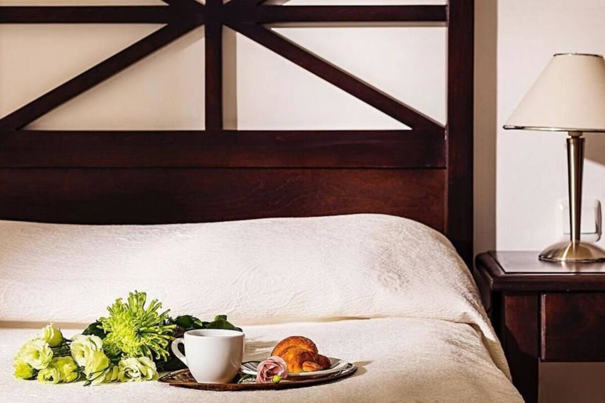 A neatly made bed with a white comforter, a tray with croissants and flowers, a white cup, and a lamp on a bedside table.