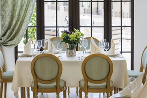 A dining table set for six with chairs, a vase of flowers, and a view through large windows. - The Cliff Beach & SPA Hotel (Obzor)