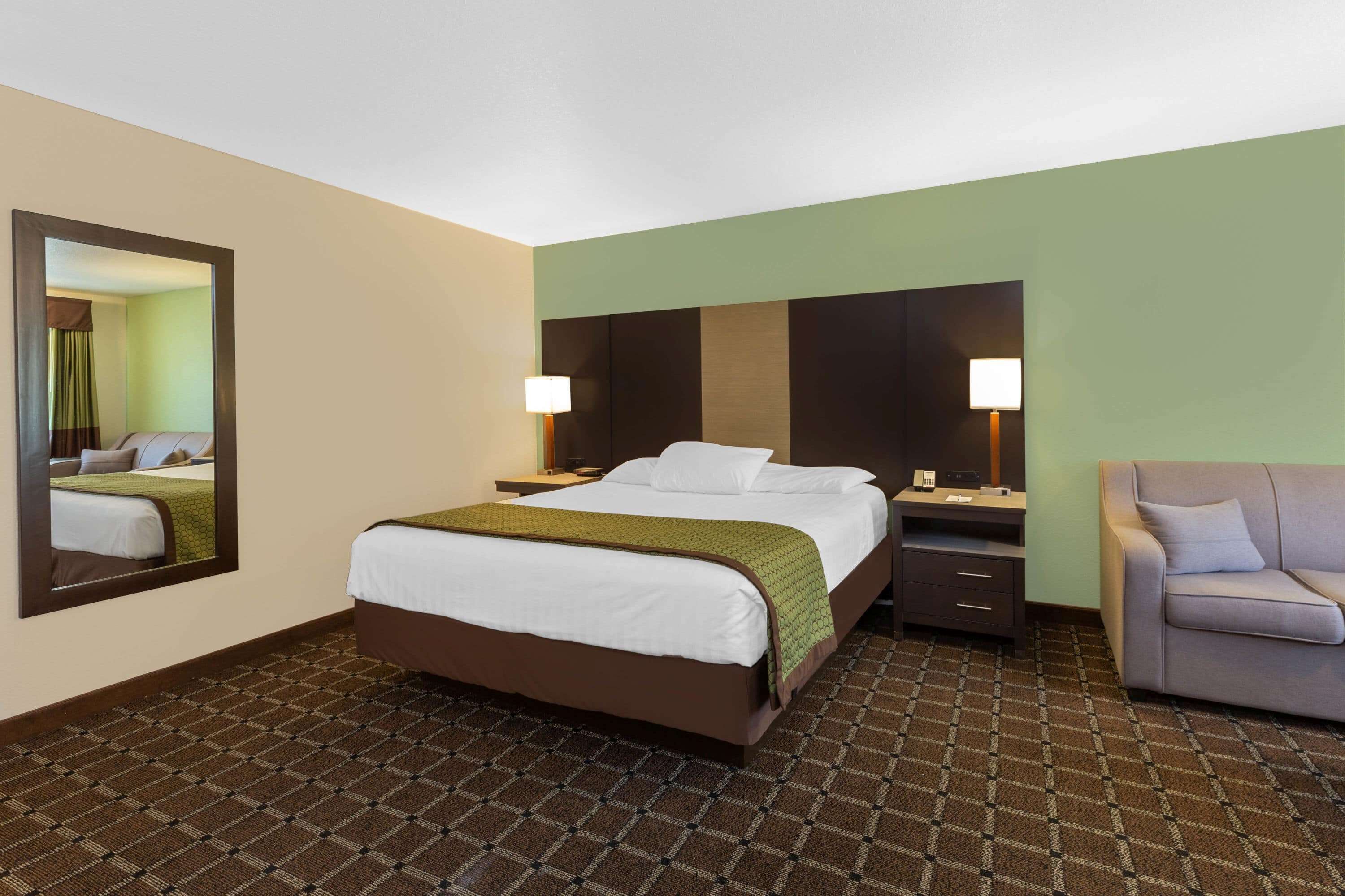 A hotel room with a bed, a desk, a chair, a mirror, and a lamp.