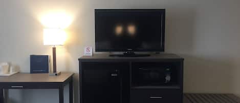 Habitación de hotel con televisor de pantalla plana sobre un mueble negro, un escritorio con lámpara, refrigerador pequeño y microondas.