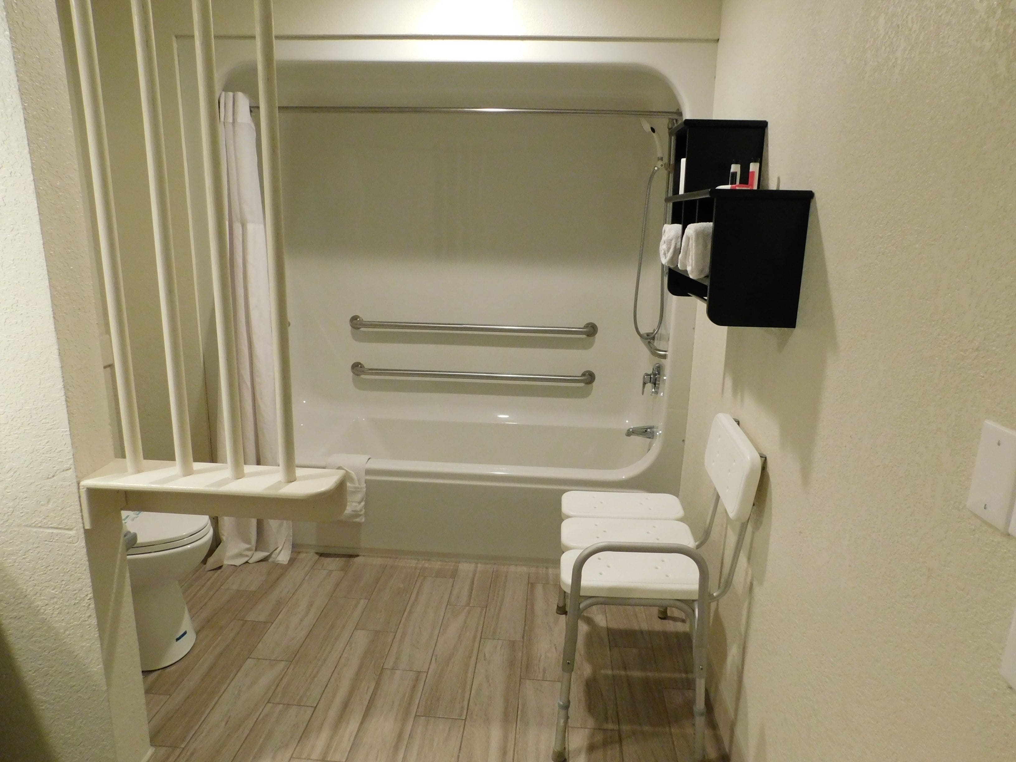 Un baño pequeño y funcional con una bañera blanca, un inodoro y una ducha. Hay dos barras de apoyo en la bañera y una pequeña silla blanca.