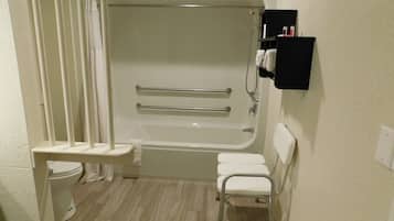 Un baño pequeño y funcional con una bañera blanca, un inodoro y una ducha. Hay dos barras de apoyo en la bañera y una pequeña silla blanca.