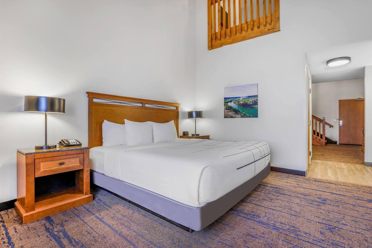 Hotelkamer met een bed met houten hoofdeinde, wit beddengoed, een nachtkastje met lamp, een tafeltje met telefoon, en een foto aan de muur.