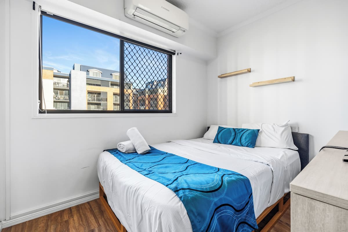A hotel room with a bed, a window with a view of buildings, a desk, and a wall-mounted air conditioner.