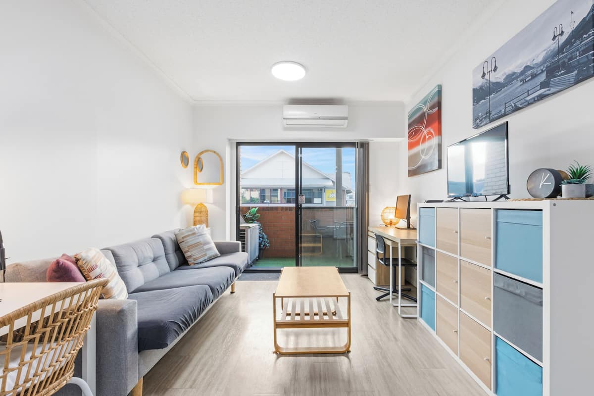 A modern living room with a sofa, a wooden coffee table, a dining area with a table and chairs, a kitchen area with cabinets, and a large window with a view of buildings.