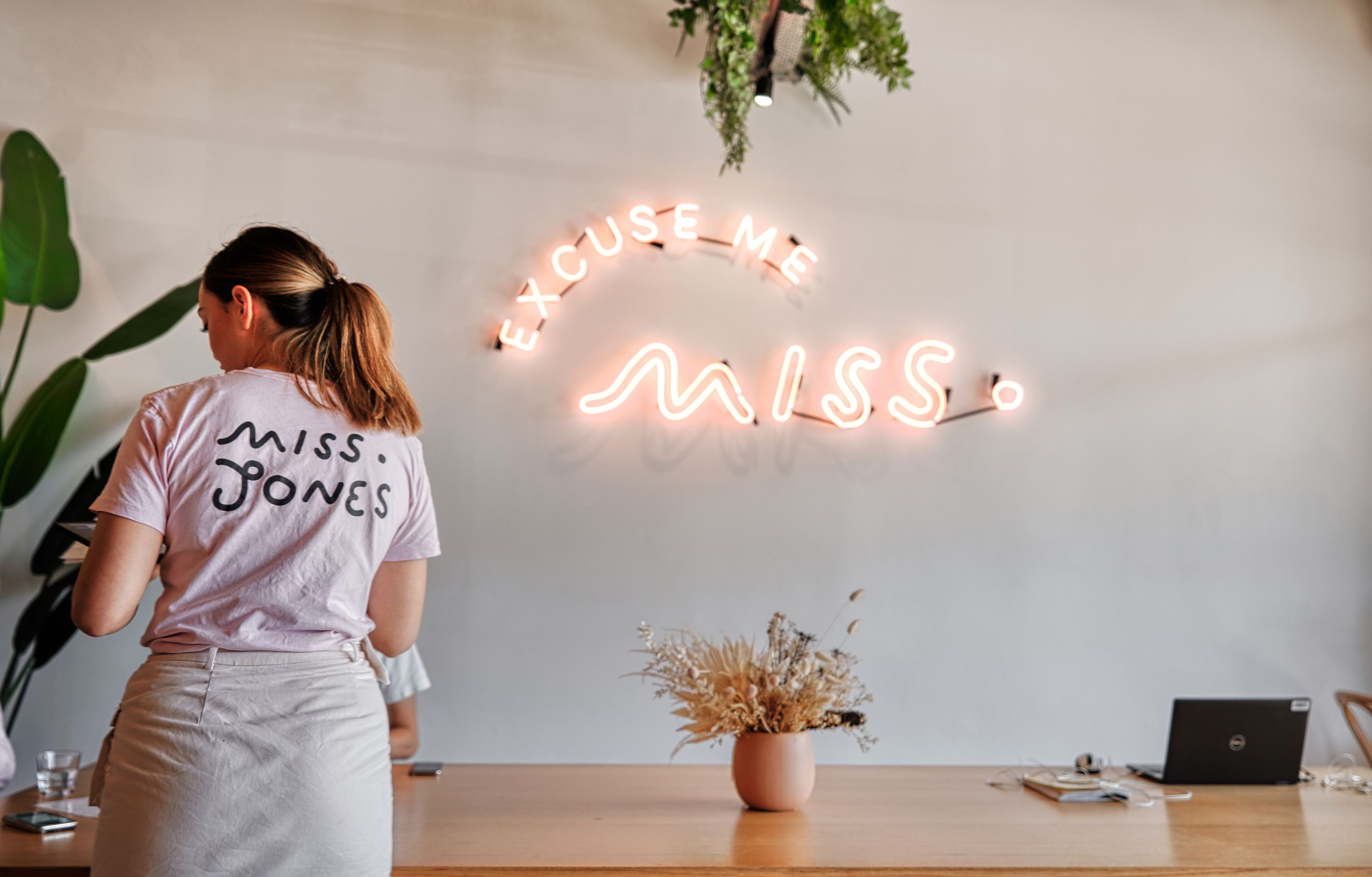 A woman in a 'Miss Jones' t-shirt standing in front of a wall with a neon sign that reads 'EXCUSE ME MISS.'