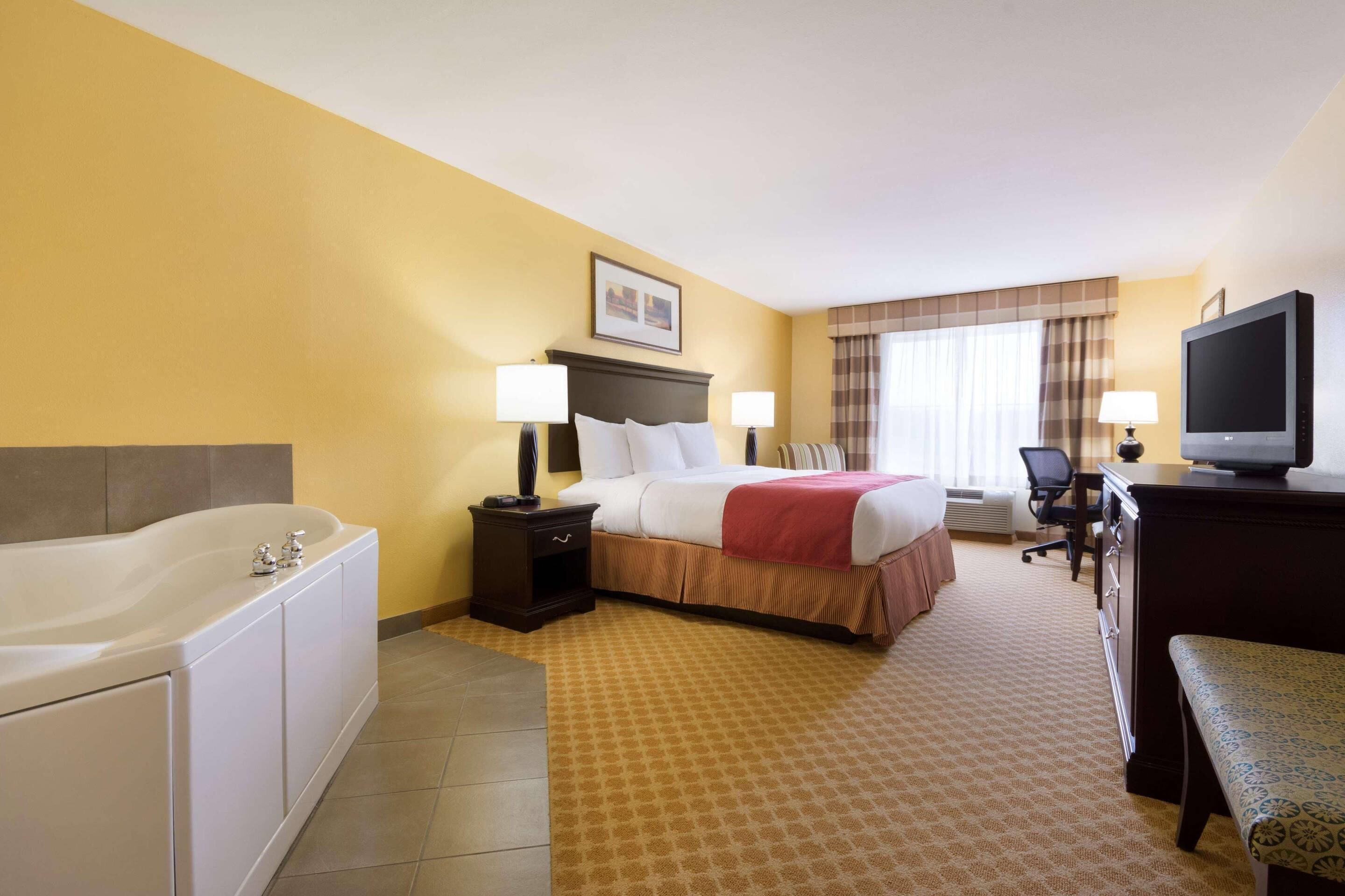 Habitación de hotel con cama, televisión, escritorio, silla, lámpara de escritorio, mesita de noche, bañera y alfombra estampada.